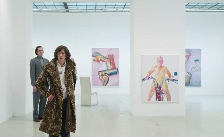 Szene aus dem Kinofilm "Mit einem Tiger schlafen" Eine Frau mit Fellmantel steht in einer Ausstellung. Im Hintergrund steht ein Mann und man sieht unscharf ein paar aufgehängte Bilder.