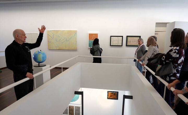 Claude Wall: Ich reite und mein Pferd geht zu Fuß – Malerei und Skulptur Künstler Claude Wall führt eine Besuchergruppe durch seine Ausstellung in der Galerie der Stadt Fellbach. Im Hintergrund sind Gemälde und ein Globus zu sehen. Foto: © Kunstverein Göppingen