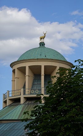 Kuppel des Kunstgebäudes am Schlossplatz in Stuttgart mit goldener Hirschskulptur, teils von Bäumen verdeckt, unter blauem Himmel mit Wolken.