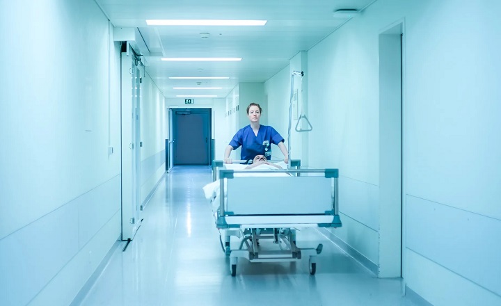 Pflegekraft im blauen Kittel schiebt ein Krankenhausbett durch einen sterilen, hell ausgeleuchteten Klinikflur. Szene aus dem Film HELDIN. Foto: © Arsenal Filmverleih