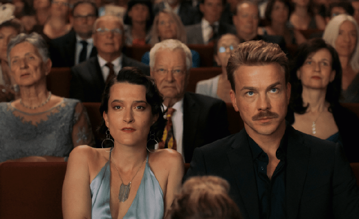 In einem prall gefüllten Theatersaal sitzt ein elegantes Paar in der ersten Reihe – die Frau trägt ein hellblaues Abendkleid, der Mann ein dunkles Hemd und einen markanten Schnurrbart. Beide blicken ernst und nachdenklich nach vorne. Hinter ihnen: ein konzentriertes Publikum, das in festlicher Kleidung einer Aufführung lauscht. Die Szene strahlt Spannung und innere Distanz aus – ein stilles Spiel der Rollen inmitten gesellschaftlicher Erwartungen.