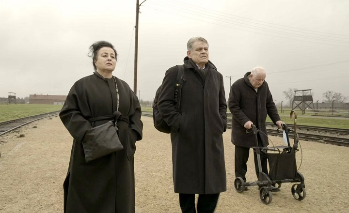 Drei ältere Menschen gehen an einem historischen Gleis entlang, das in ein ehemaliges Konzentrationslager führt. Alle tragen dunkle Mäntel; zwei von ihnen laufen aufrecht, der Dritte stützt sich auf einen Rollator. Die Stimmung ist still und nachdenklich, der Himmel grau. Im Hintergrund sind Wachtürme und Baracken zu erkennen – ein Ort der Erinnerung und des Schmerzes.