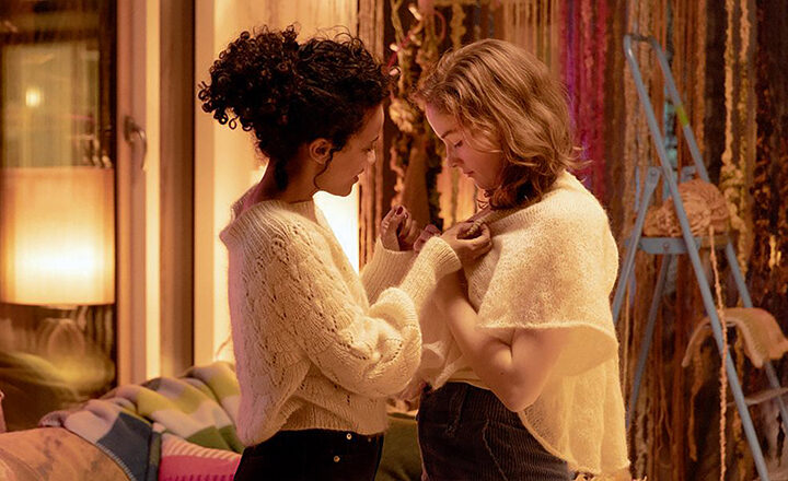 Zwei junge Frauen stehen sich in einem warm beleuchteten Raum gegenüber. Beide tragen kuschelige, helle Strickpullover. Sanft und behutsam richtet die eine den Kragen der anderen, während ihre Blicke sich in inniger Zärtlichkeit begegnen. Im Hintergrund sorgen bunte Textilien und weiches Licht für eine intime, geschützte Atmosphäre.
