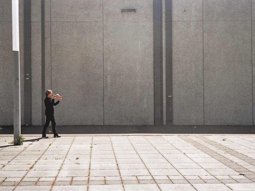 Die Konzeptkünstlerin Kathleen Jahn steht auf einem leeren, gepflasterten Platz vor einer großen grauen Betonwand und spielt ein Schneckenhorn. Der Ort wirkt reduziert und klanglich offen.
