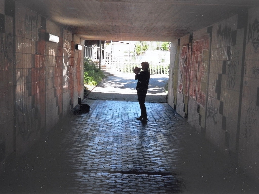 Kathleen Jahn steht allein in einer graffitibeschmierten Unterführung und bläst in ein Schneckenhorn. Tageslicht fällt vom Ausgang auf den Pflasterboden und verstärkt die akustische Atmosphäre des Raums.