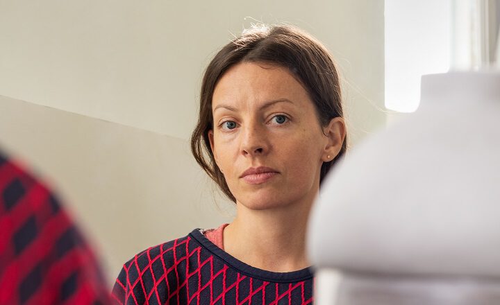 Porträt der Künstlerin Anna Lea Hucht. Sie blickt ruhig in die Kamera, trägt einen dunkelblauen Pullover mit rotem Rautenmuster. Im Vordergrund ist unscharf ein weißes Objekt zu sehen.