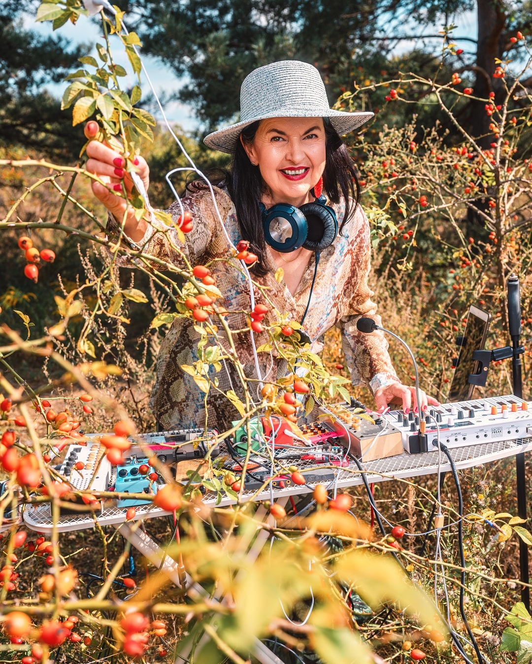 Cassis B Staudt Klangkünstlerin Cassis Birgit Staudt steht inmitten von Sträuchern mit Hagebutten und spielt an einem elektronischen Musiksetup; Elektroden sind an Pflanzen befestigt.