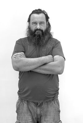 Schwarz-weißes Porträt eines Mannes mit langem Bart und zusammengebundenen Haaren, der mit verschränkten Armen vor einer weißen Wand steht.