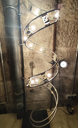 Lichtinstallation von Walter Giers im Schmiedturm in Schwäbisch Gmünd: Eine spiralförmige Skulptur mit leuchtenden Glühbirnen und elektronischen Bauteilen vor einer rauen Steinwand.