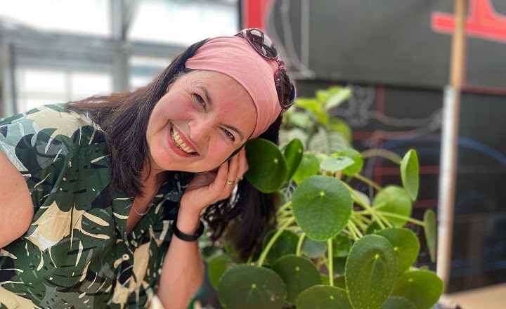 Klangkünstlerin Cassis Birgit Staudt lehnt lächelnd an einer Pflanze und hört deren Signale; im Hintergrund ein Innenraum.