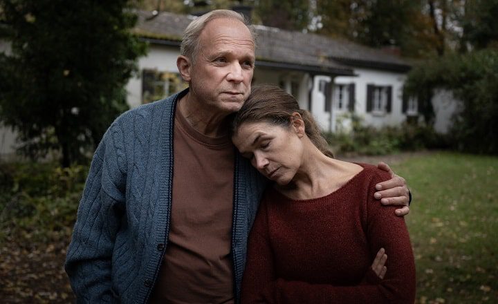 Filmszene aus Dann passiert das Leben: Ein Mann und eine Frau stehen eng beieinander im Freien, die Frau lehnt erschöpft ihren Kopf an die Schulter des Mannes, der sie tröstend im Arm hält.