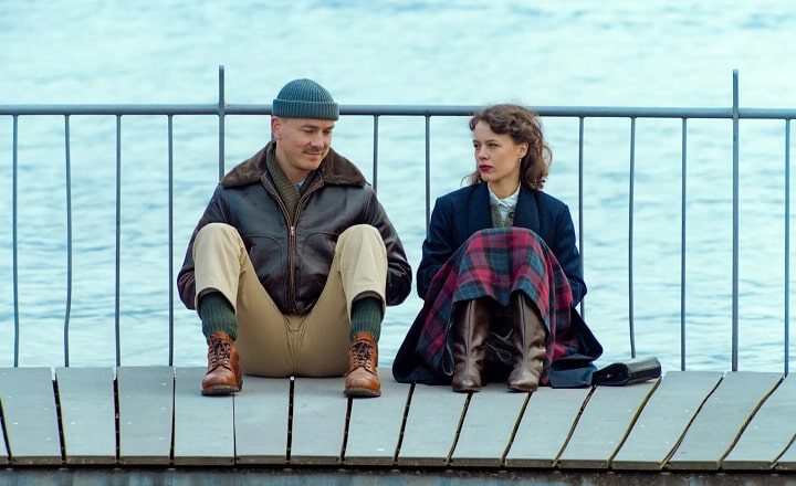 Filmszene aus Stiller: Ein Mann und eine Frau sitzen nebeneinander auf einem Holzsteg am Wasser, getrennt durch Abstand und Blickrichtung, in ruhiger, nachdenklicher Stimmung.