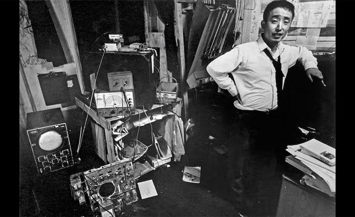 K3 Programmhinweis: NAM JUNE PAIK: MOON IS THE OLDEST TV Schwarz-weiße Archivaufnahme von Nam June Paik in einem Atelier: Der Künstler steht zwischen elektronischen Geräten, Kabeln und Monitoren und blickt in die Kamera.