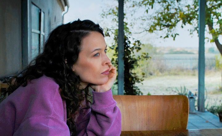 Filmszene aus Miroirs No. 3: Eine Frau sitzt nachdenklich auf einer Bank, den Kopf in die Hand gestützt, und blickt in die Landschaft vor einem Haus.