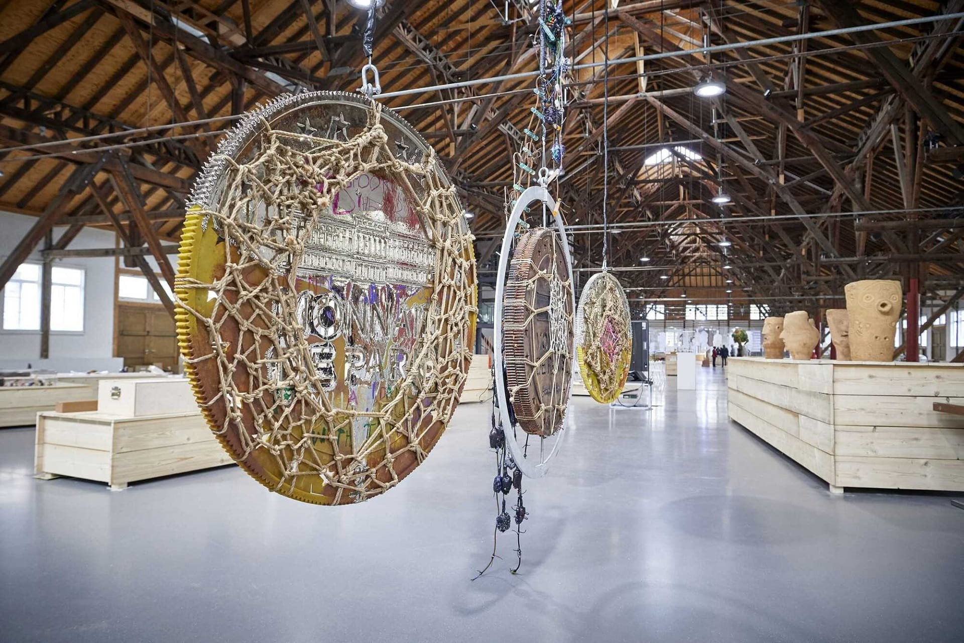 kaya_kaya_moo_nay_moon_ay_moonay_triennale_fellbach_peter_d_hartung_2019 Hängende, kreisförmige Skulpturen aus verschiedenen Materialien in der Ausstellungshalle der Triennale Fellbach; im Hintergrund weitere Werke und die historische Architektur der Alten Kelter.