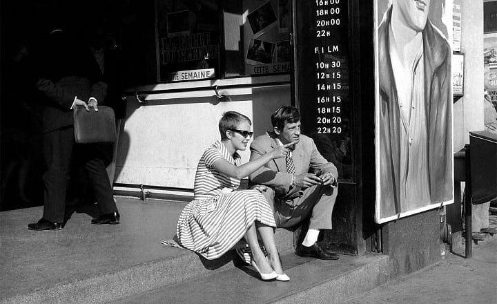 Ein Schwarzweißbild zeigt eine Frau mit Sonnenbrille und gestreiftem Kleid sowie einen Mann im Anzug, die gemeinsam vor einem Kinoeingang auf einer Stufe sitzen.