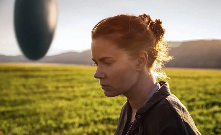 Filmstill aus „Arrival“: Eine Frau steht nachdenklich auf einer Wiese, im Hintergrund schwebt eines der fremdartigen Raumschiffe.