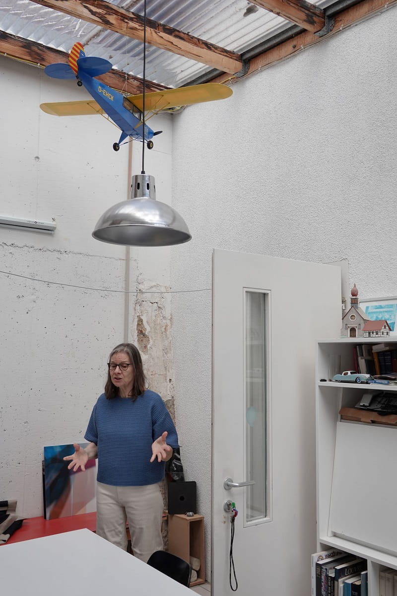 Simone Demandt spricht in ihrem Atelier in Baden-Baden-Varnhalt zur Exkursionsgruppe des Kunstvereins Göppingen. Über ihr hängen eine silberne Lampe und ein blau-gelbes Modellflugzeug.