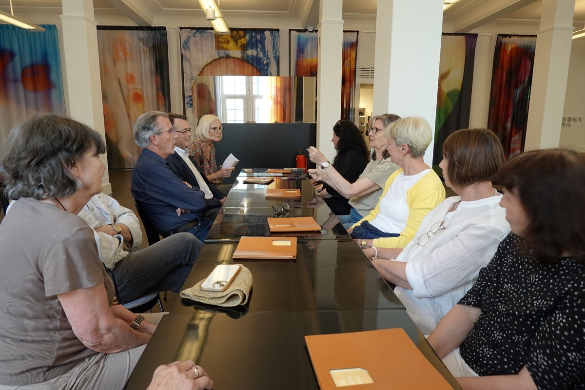 Die Exkursionsgruppe des Kunstvereins Göppingen sitzt in Baden-Baden an einem langen Tisch, umgeben von großformatigen farbigen Arbeiten.