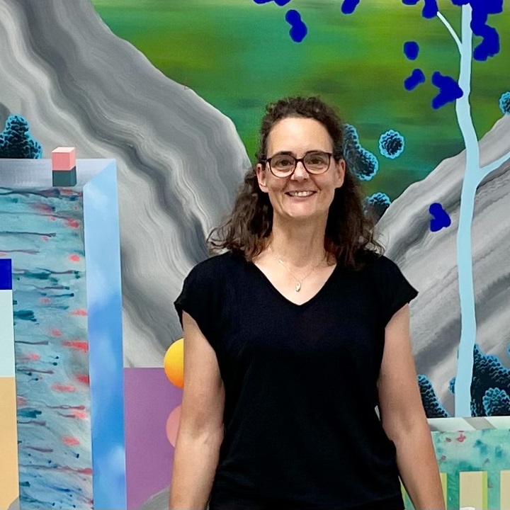 Eine Frau steht lächelnd vor einem farbenfrohen Kunstwerk von Leo Staigle in Grün-, Blau-, Grau- und Rosatönen.