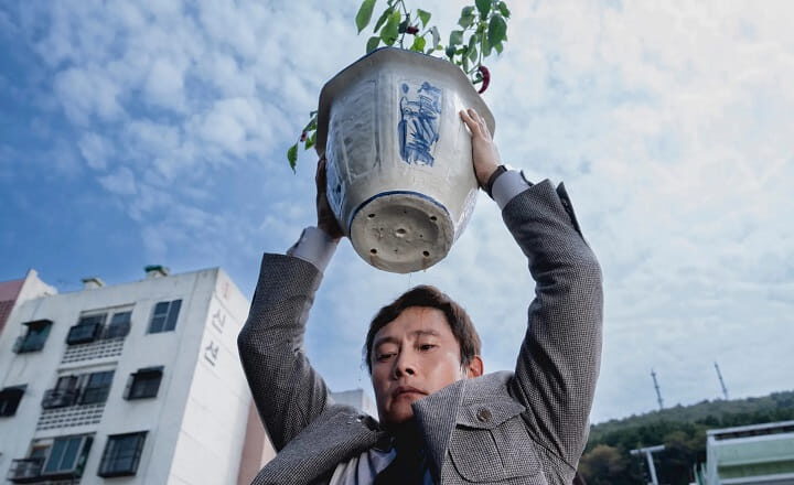 Filmszene aus „No Other Choice“: Ein Mann trägt einen großen Blumentopf über dem Kopf.
