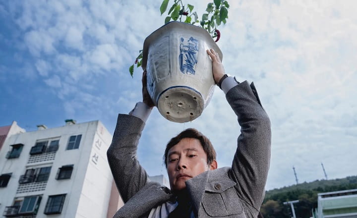 Filmszene aus „No Other Choice“: Ein Mann trägt einen großen Blumentopf über dem Kopf.